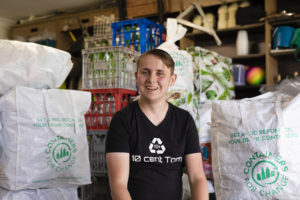 A man wearing a black t-shirt with the writing 10 Cent Tom sits in front of bags of drink containers.