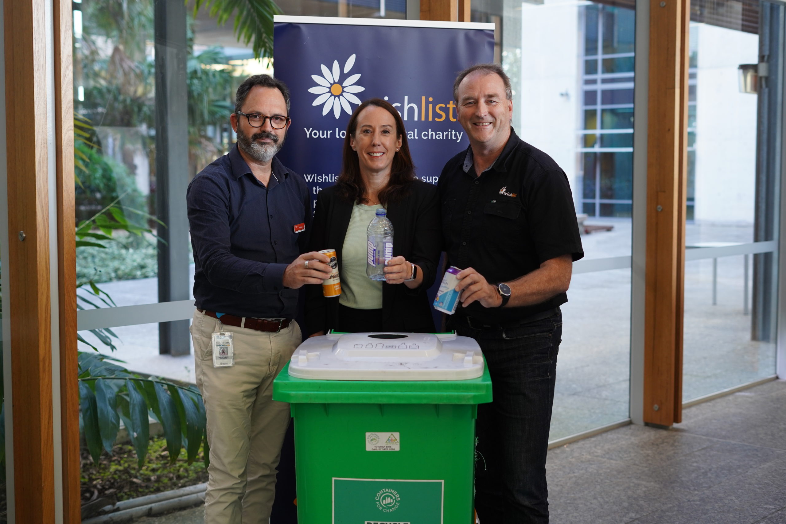 Three people hold containers above a green recycling bin.