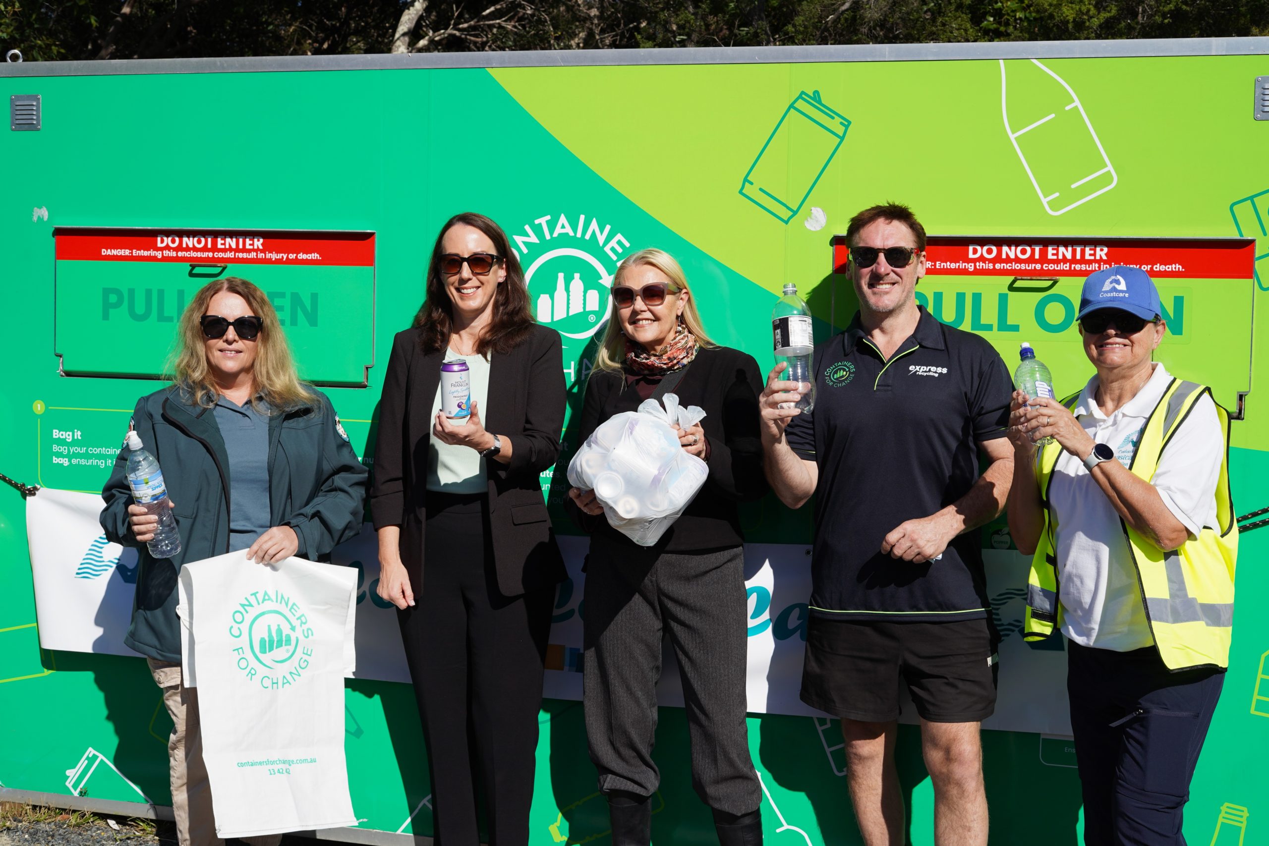 Five people stand in front of a green shipping container sized bin while holding bottles and cans.