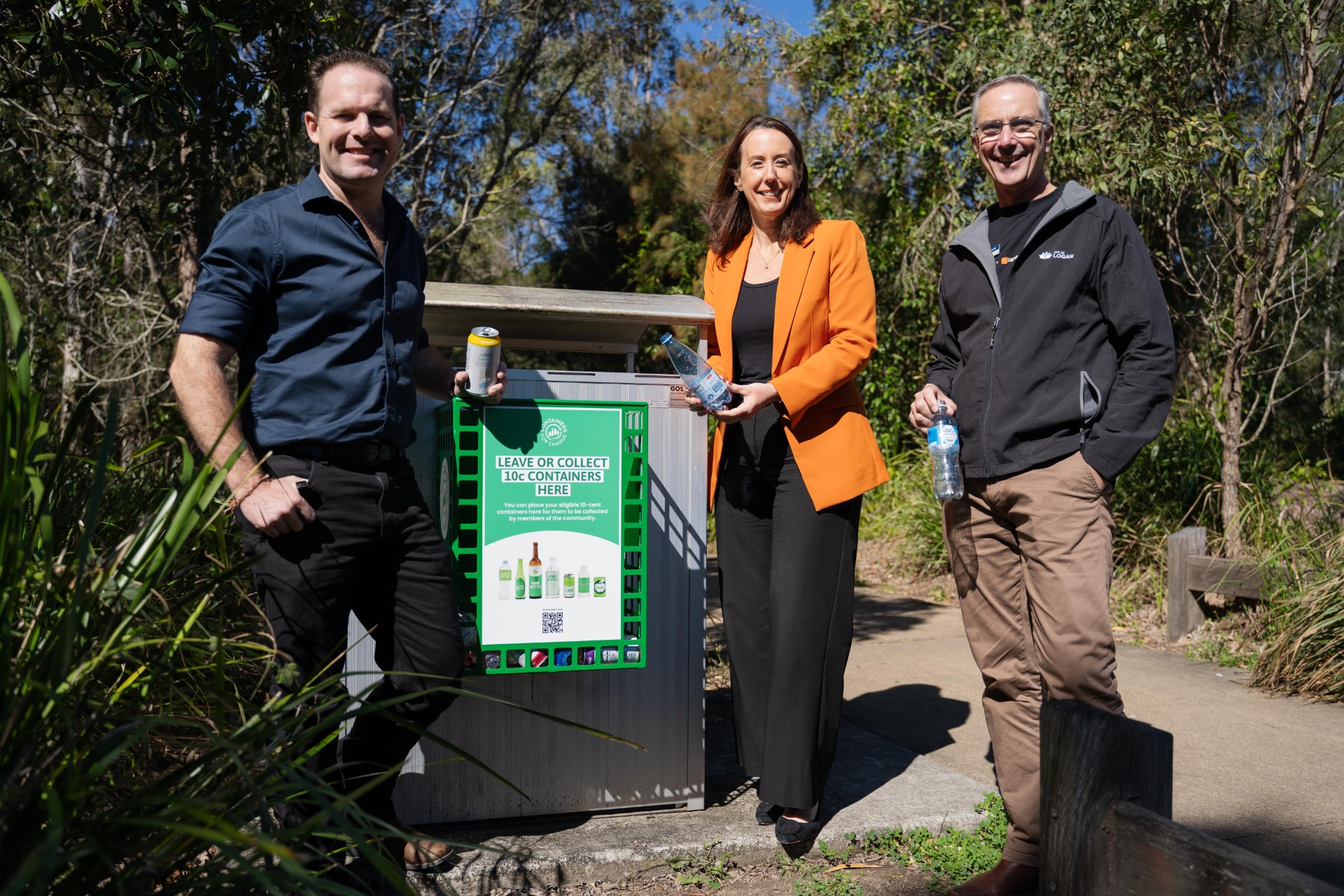 Three people holding drink containers stand next to a bin with a large green basket designed to collect drink containers attached to it.