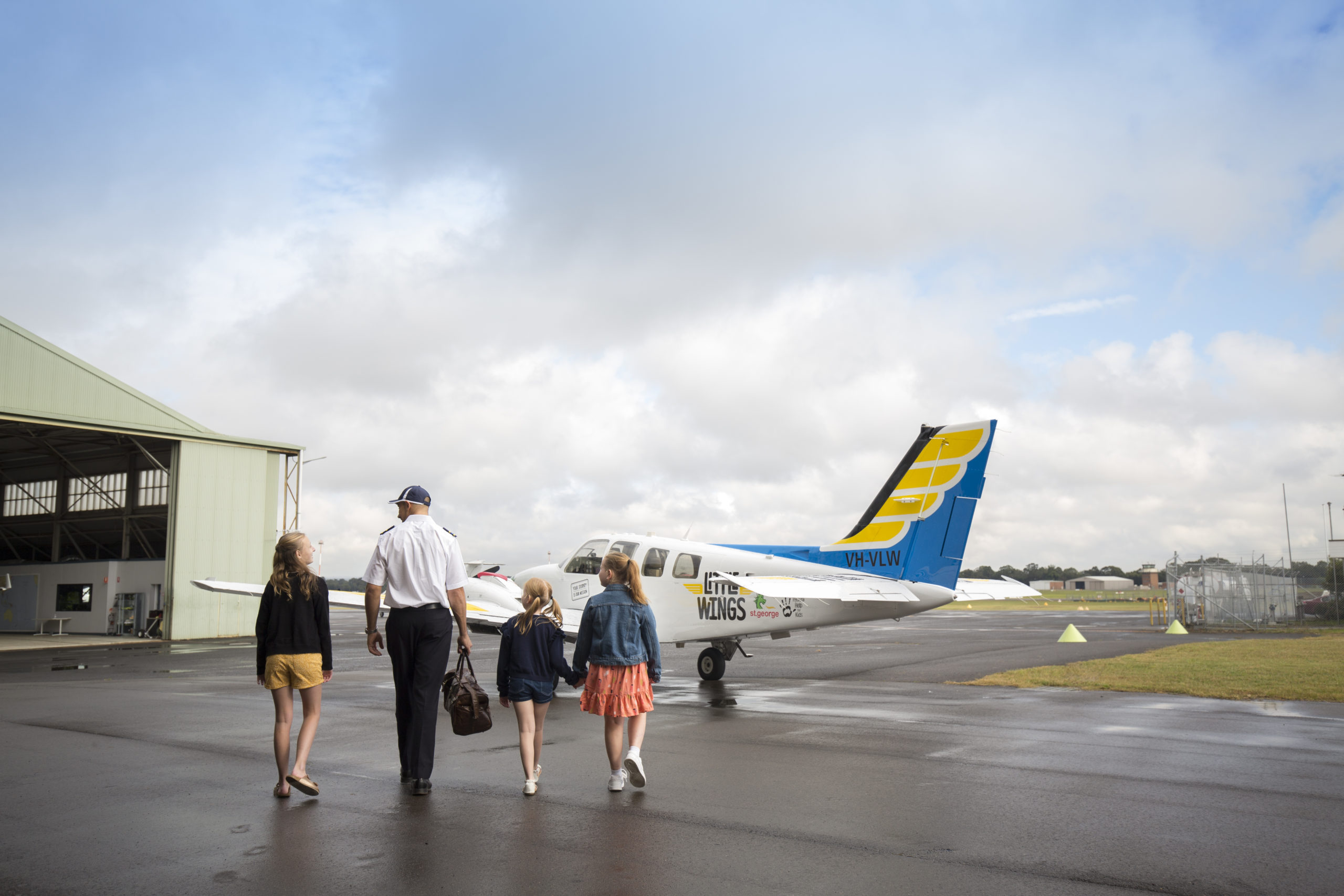 A family and a pilot walk towards a Little Wings light aircraft on the tarmac of an airport.