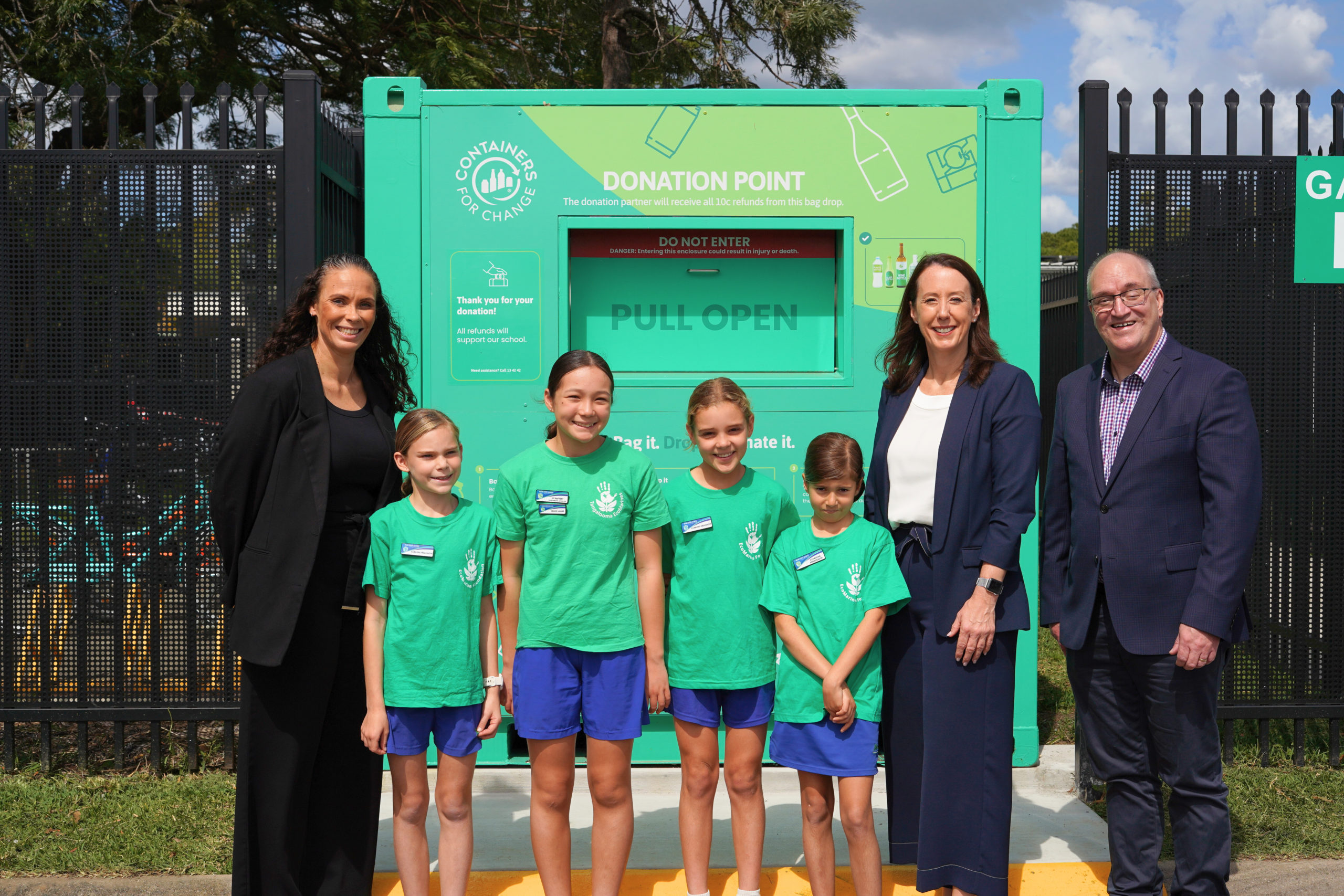 A group of adults and primary-school aged children stand in front of a large green shipping container with the words 'donation point' written on a shute.