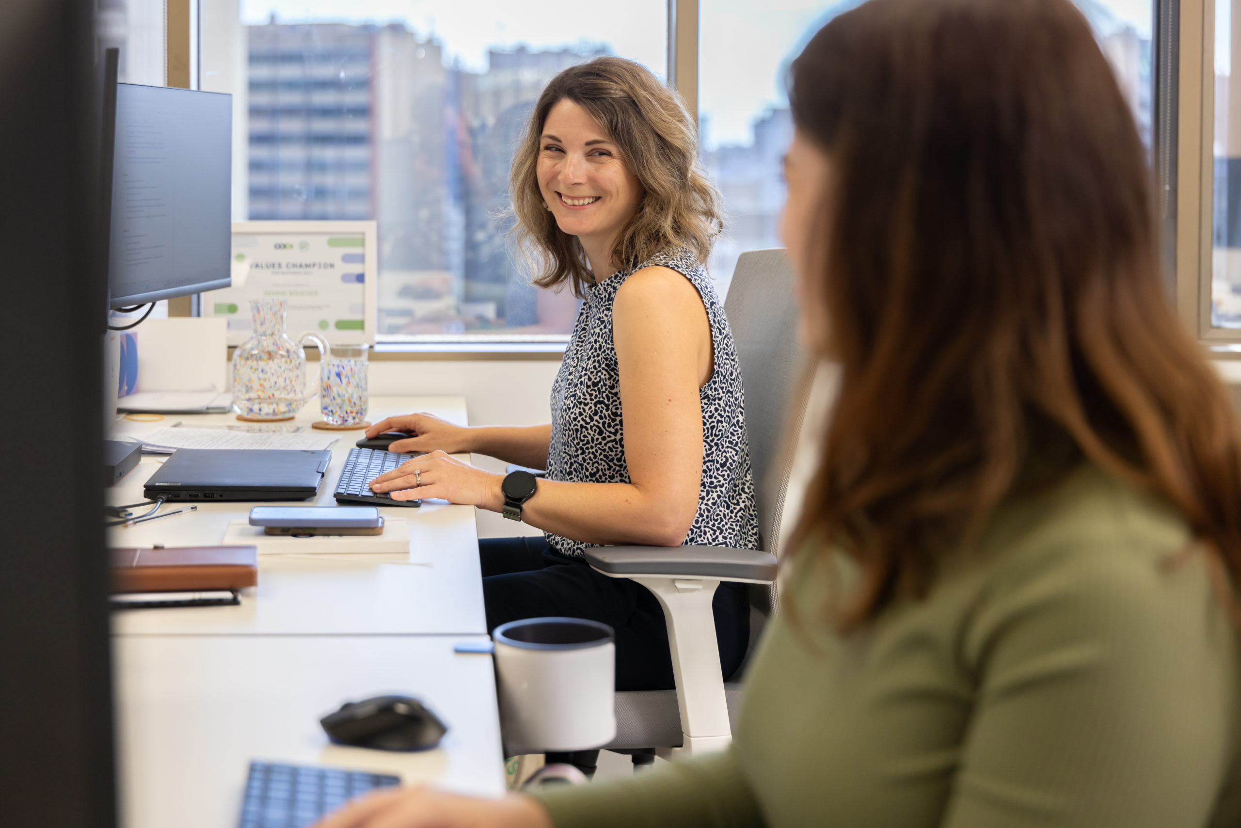 A woman sitting at a computer desk smiles at the person next to her.