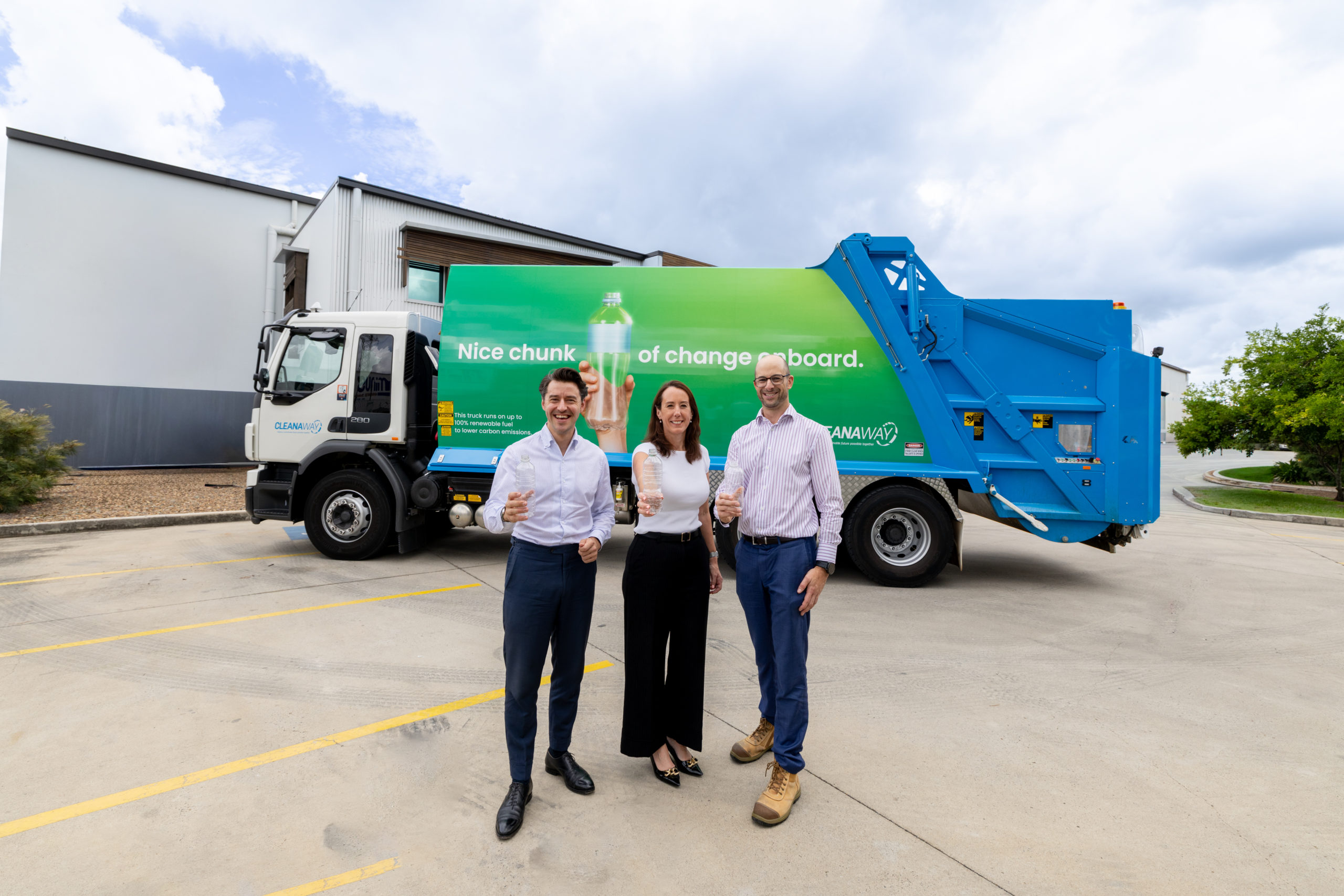 Two men and a woman smile and hold clear empty plastic bottles while standing in front of a large truck that collects empty drink containers and runs on renewable diesel.