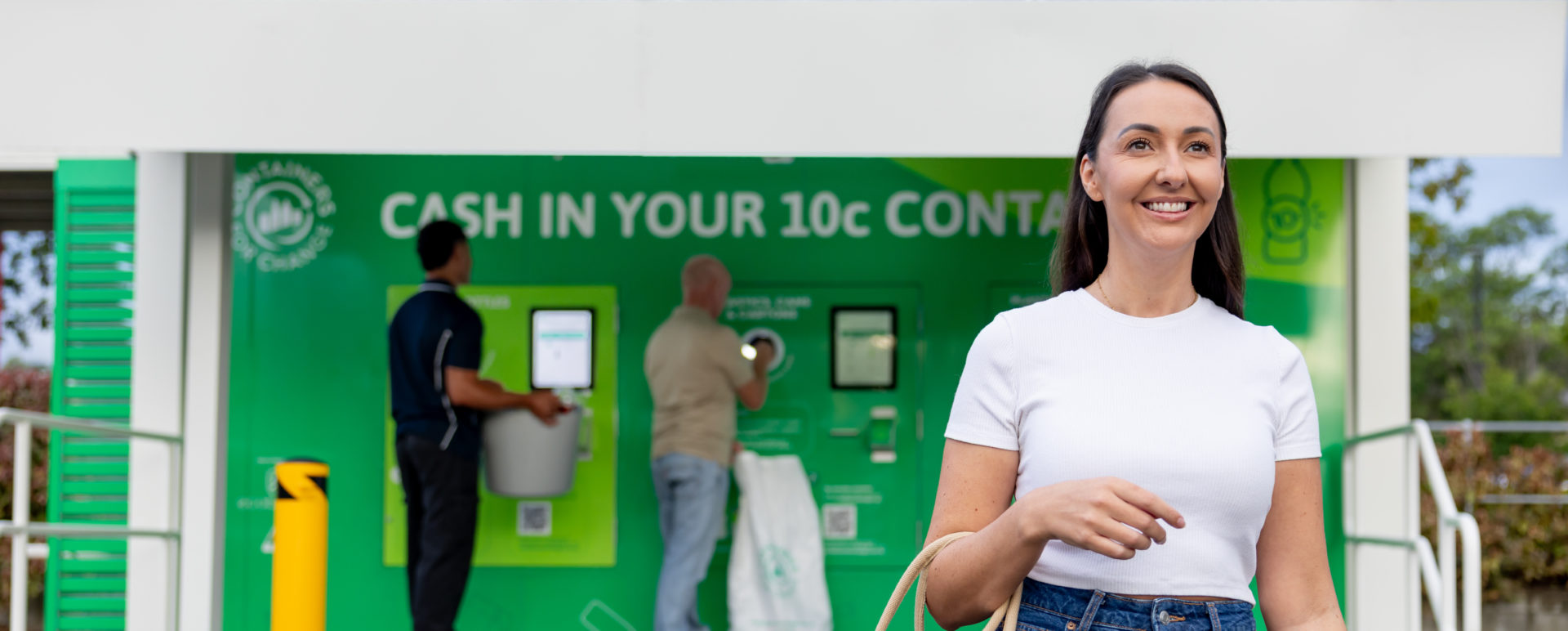 A woman holding a bag walks in front of a large green containers for change reverse vending machine.