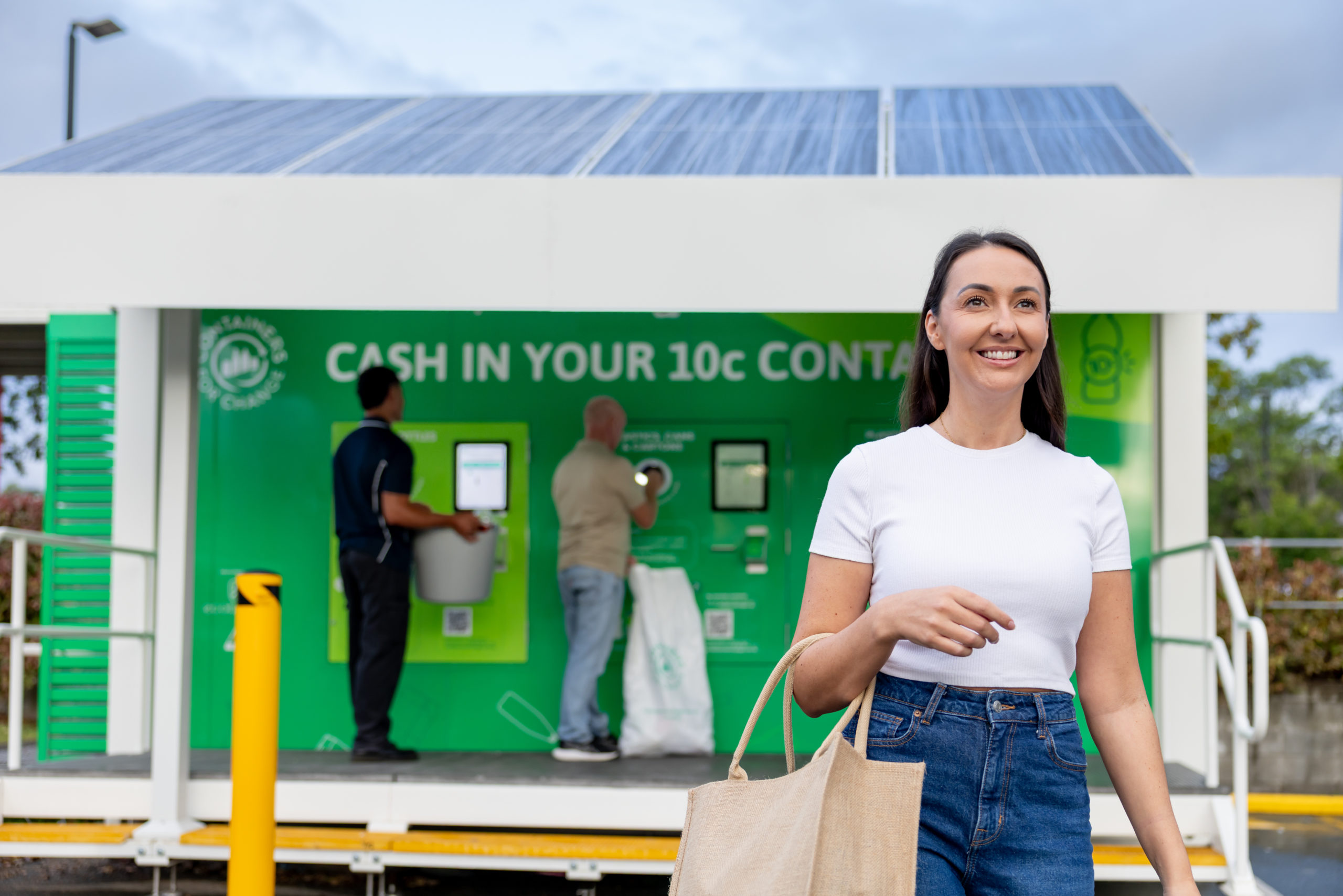 A woman holding a bag walks in front of a large green containers for change reverse vending machine.