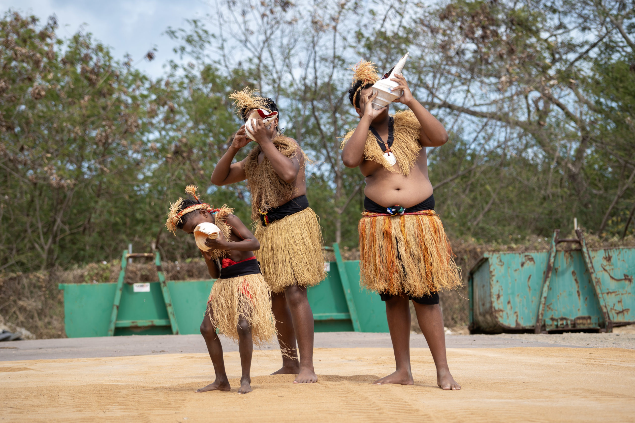 Two young men and a boy dressed in traditional grass skirts and blowing into conch shells.
