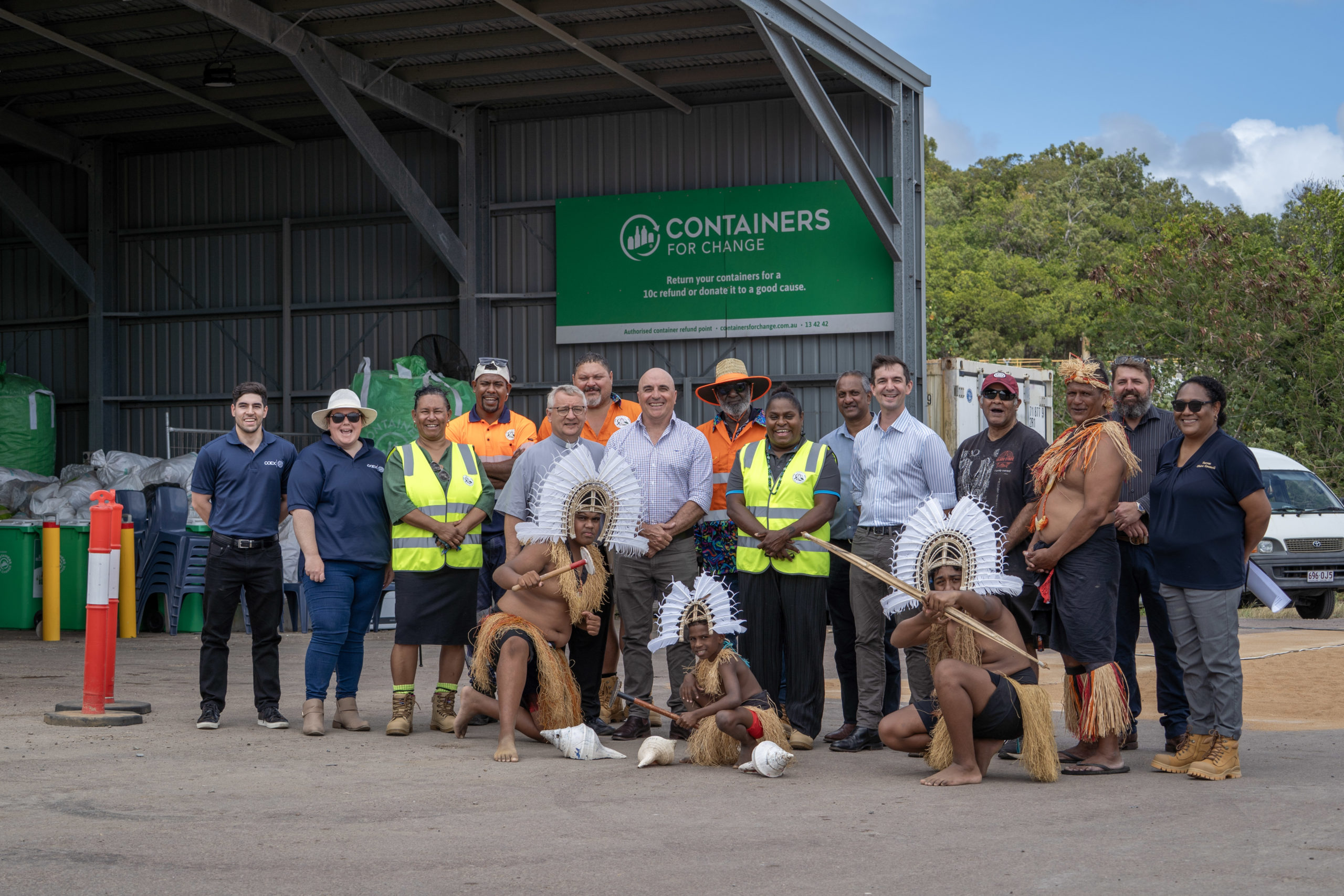 A group of people smile at the camera as they stand infront of a large recycling facilility.
