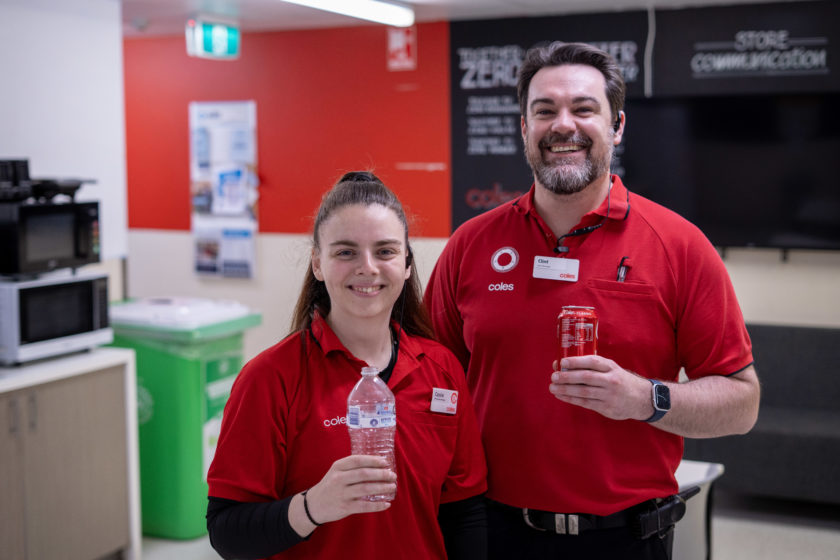 Two Coles staff members in red uniforms holding empty drink containers beside a green recycling bin.