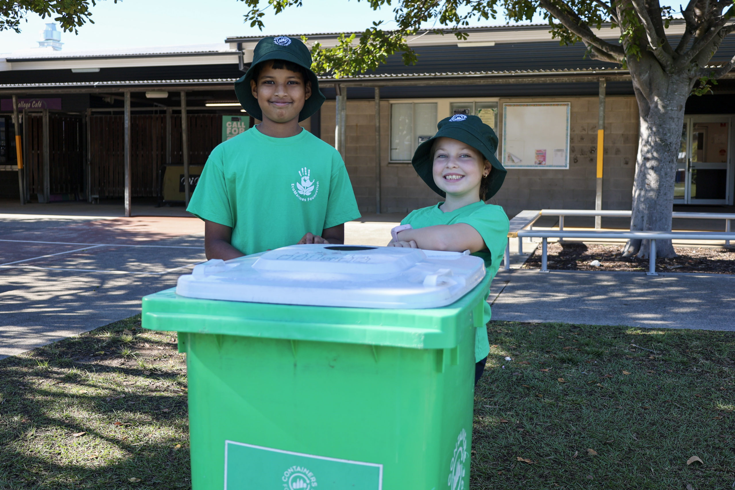 Two students standing behind a green recycling bin in a schoolyard.