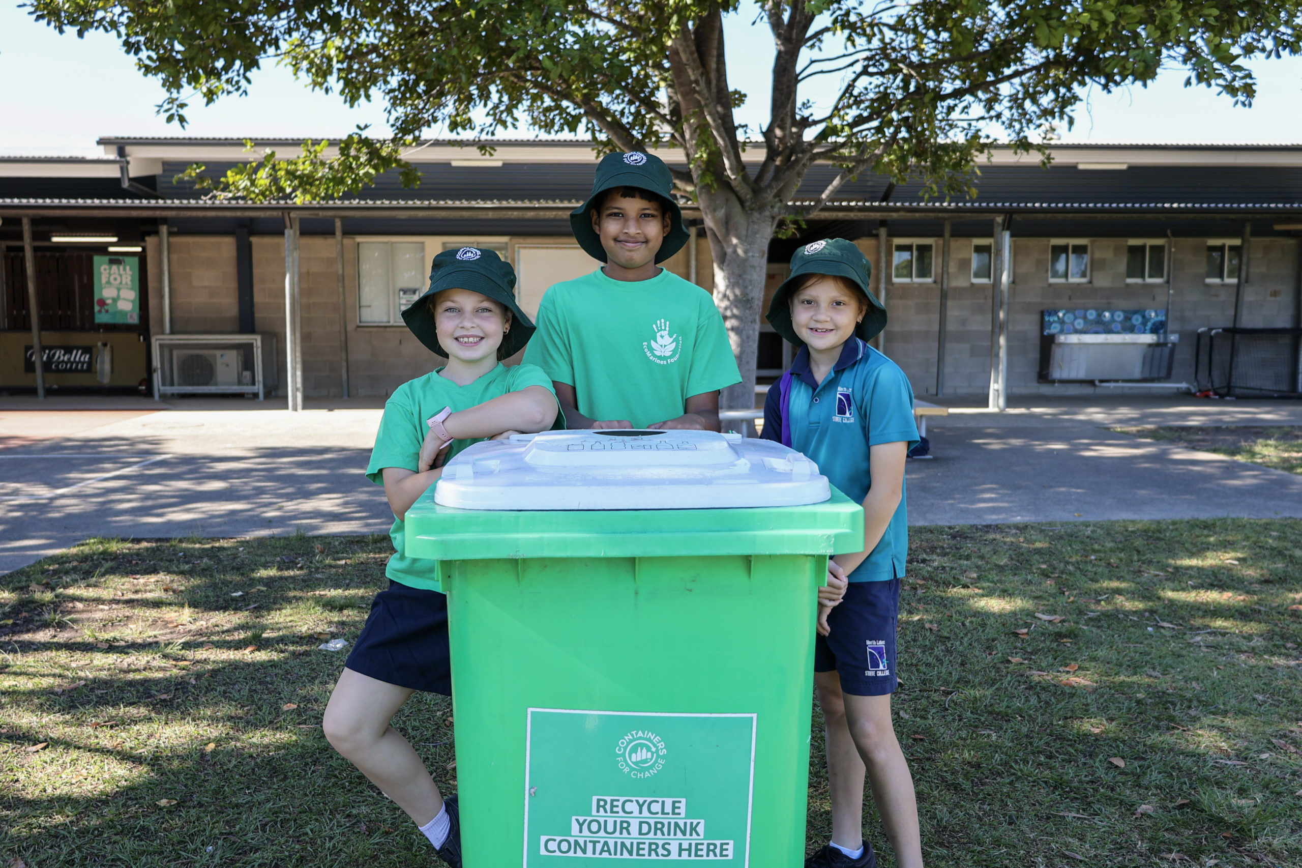 Three students standing behind a green container recycling bin in a schoolyard.