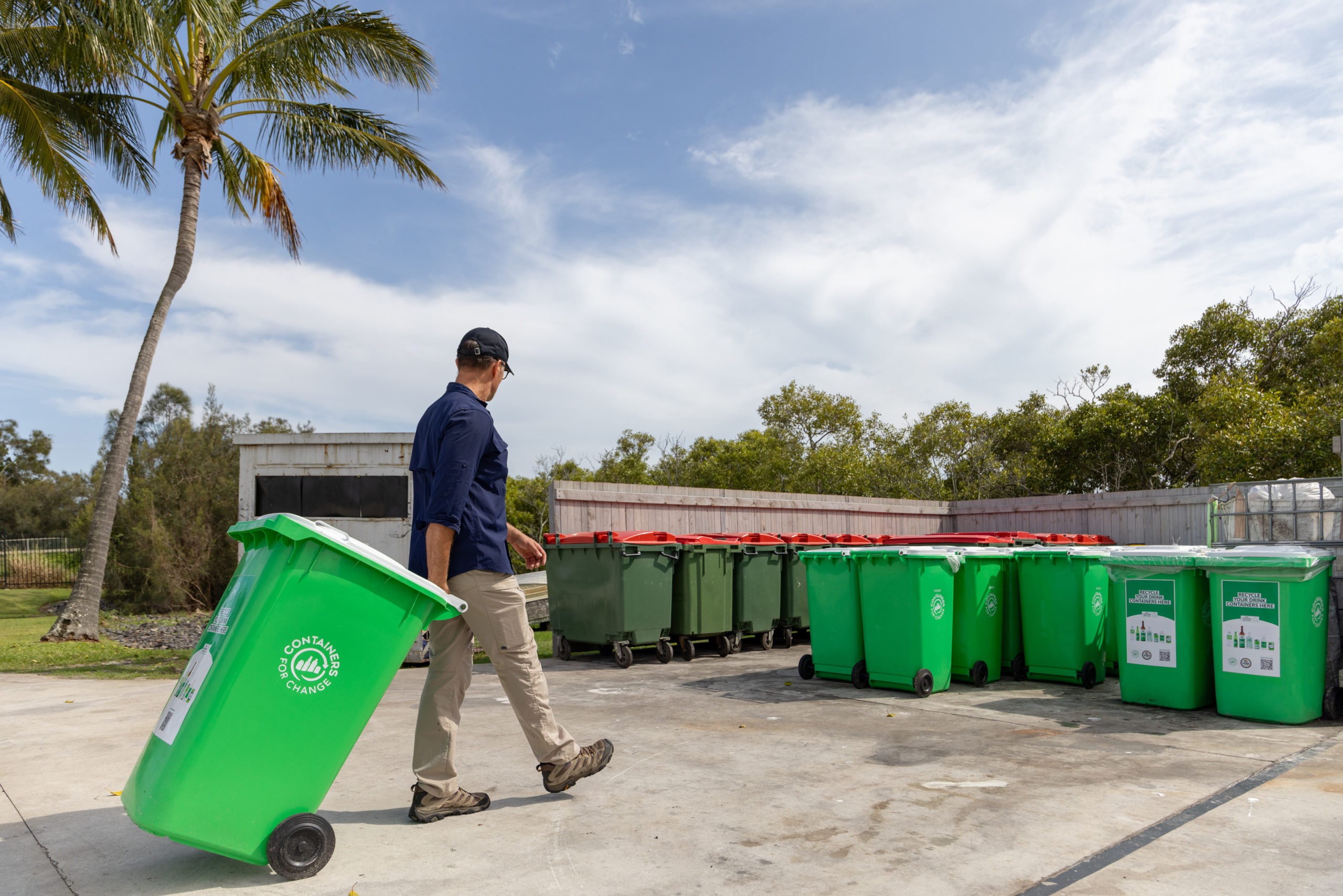 Person wheeling a green recycling bin towards a row of green collection bins at an outdoor facility.