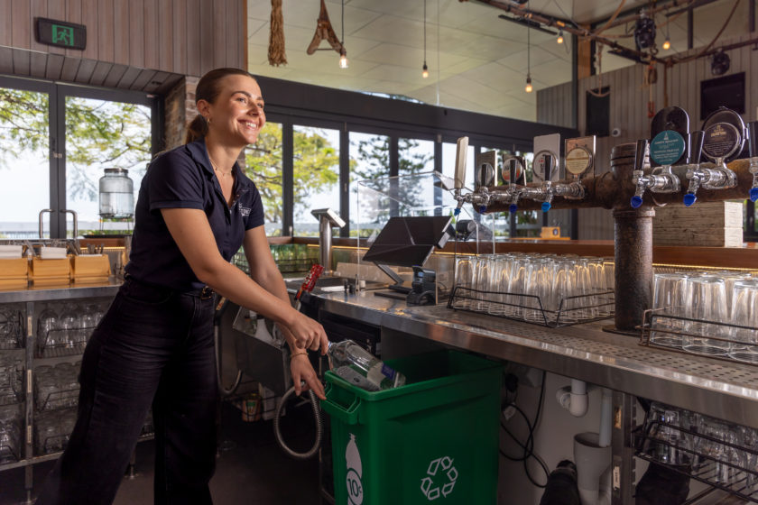 A bartender behind a bar putting an empty glass spirits bottle into a green Containers for Change bin underneath the bar, with a 10-cent logo on the side of the bin.