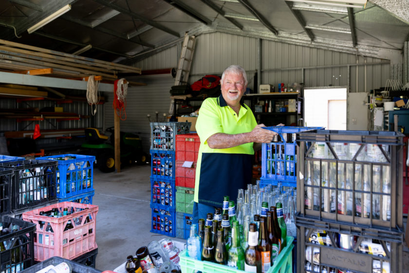 Bruce Ferguson in a high-vis yellow shirt stands inside a shed surrounded by crates filled with empty bottles and cans, with more containers stacked in the foreground.