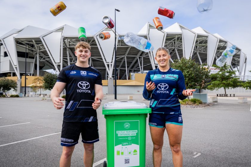 Cowboys players Xavier Kerrisk and Makenzie Weale standing beside a green Containers for Change recycling bin outside Queensland Country Bank Stadium.