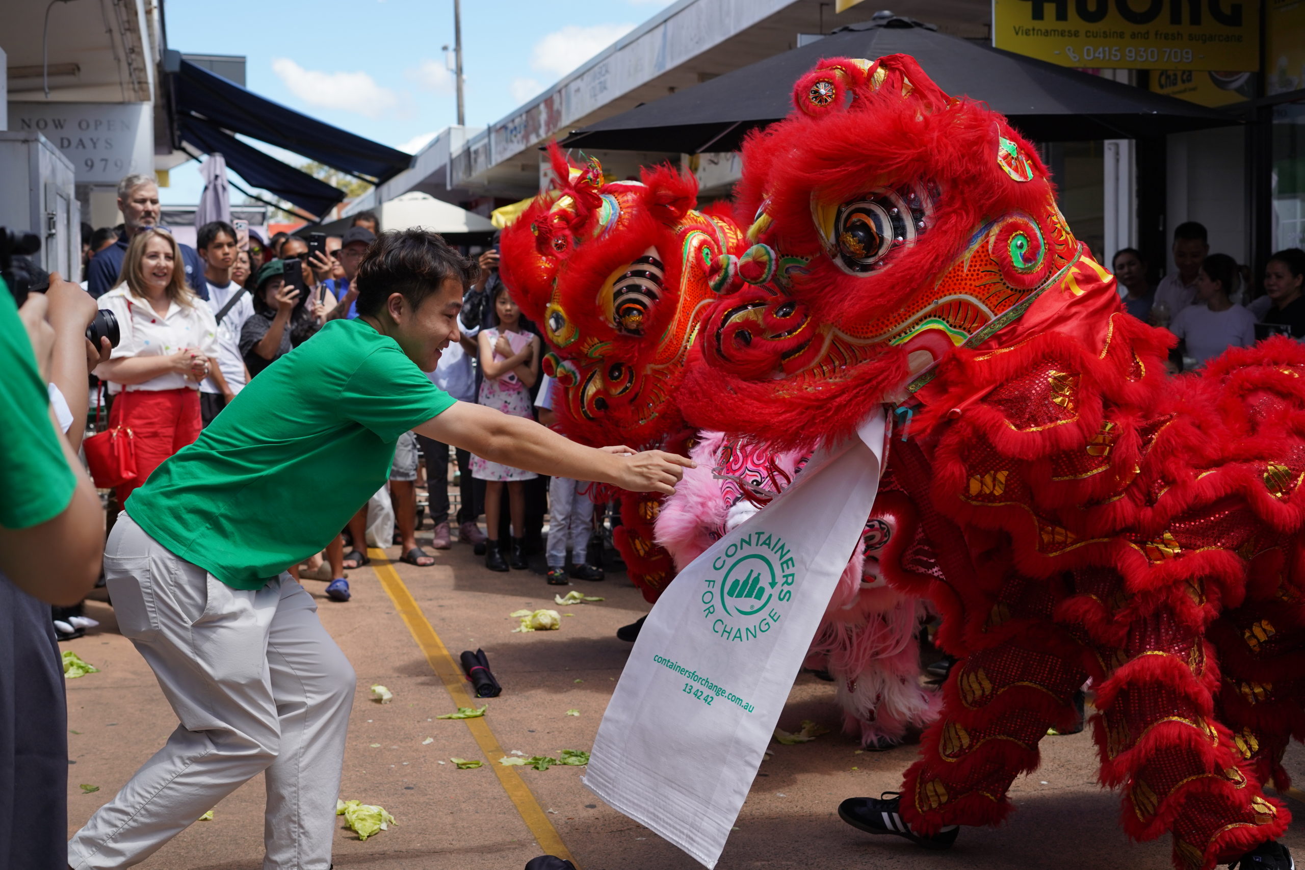 A man hands a white Containers for Change bag to a dancing lion.
