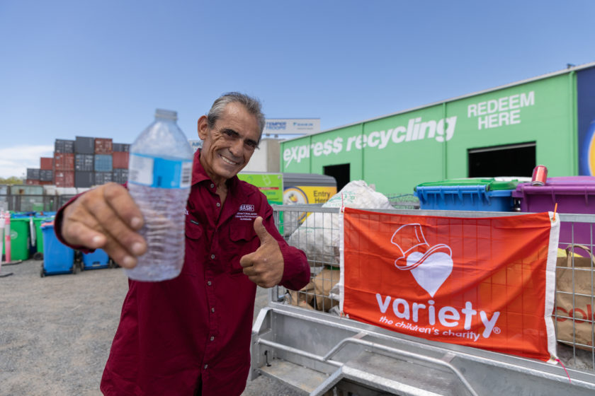 Paul Quarrell holds up an empty plastic bottle beside a container collection bin displaying the Variety logo, with a recycling facility in the background.